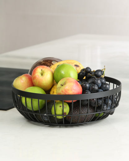 WarmyNest round wire fruit basket in matte black on a marble kitchen bench, filled with apples, limes, grapes, bananas and avocados