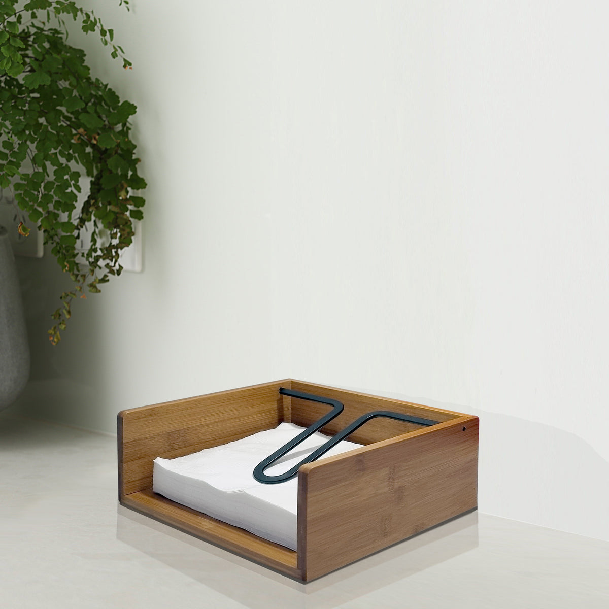 Bamboo napkin and serviette holder on a kitchen bench, filled with white napkins held down by the black metal arm, beside a potted fern